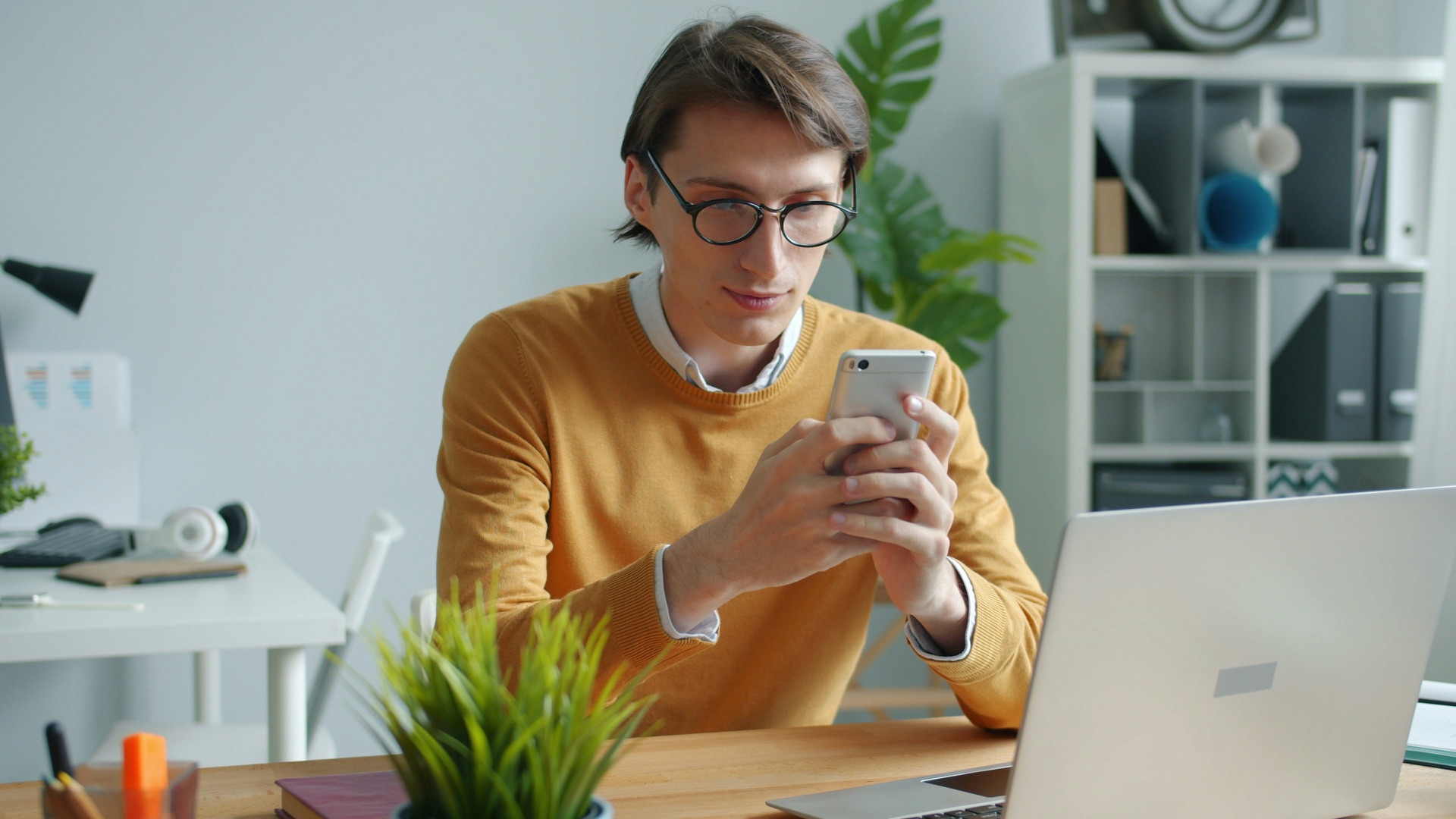 A man in glasses typing on his smartphone at an office desk, with an open laptop in the foreground.