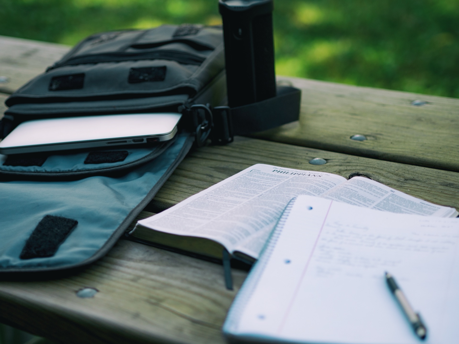 A student’s open notebook with handwritten notes and a pen, next to an open textbook and a laptop in a bag on a wooden outdoor table.
