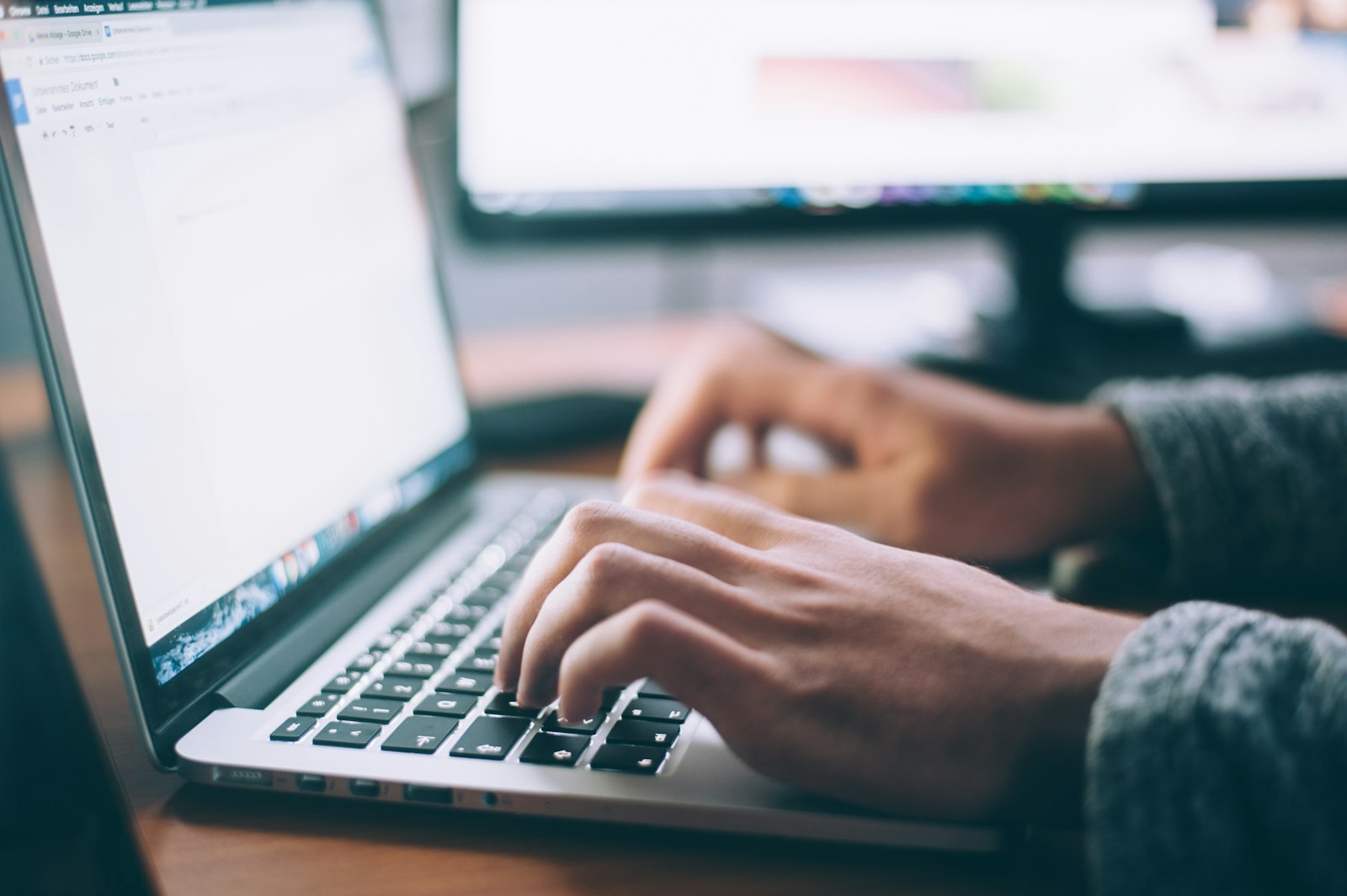 Hands typing on a laptop keyboard with a second monitor in the background.