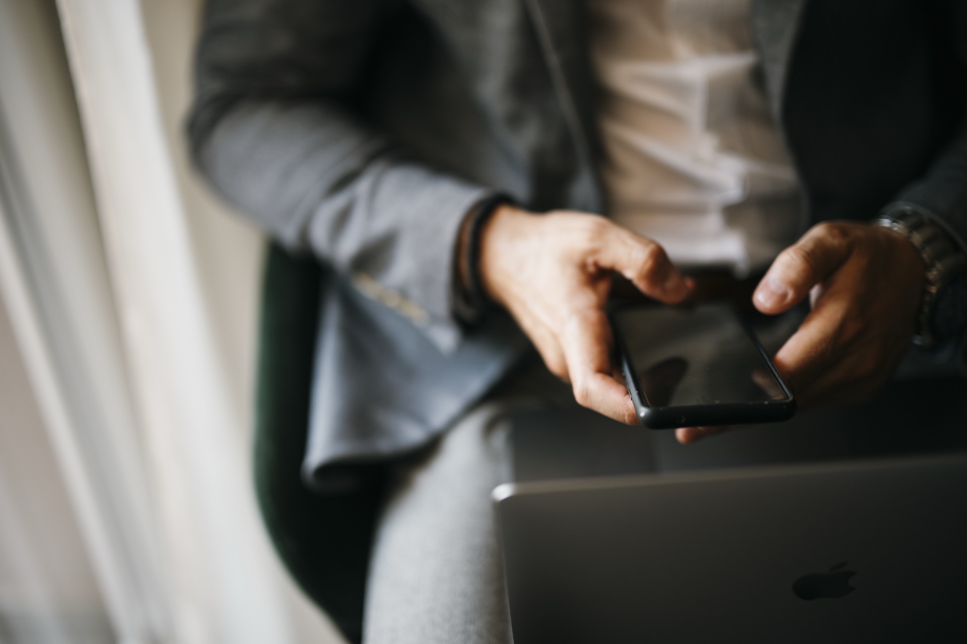 A person in a grey blazer holding a smartphone in both hands, with a laptop resting on their lap.