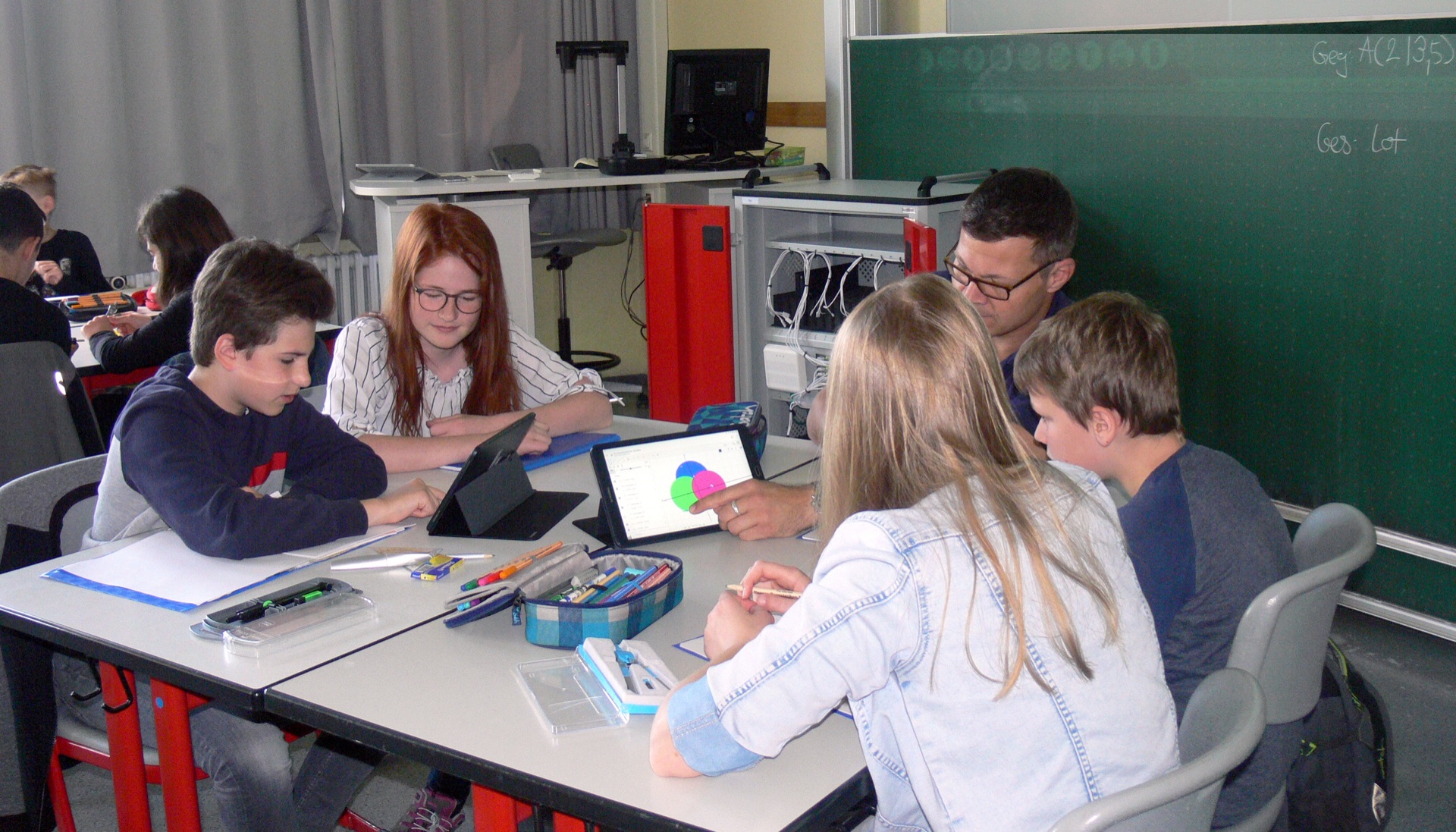 Students working on tablets around a classroom table while a teacher points at one of the screens.