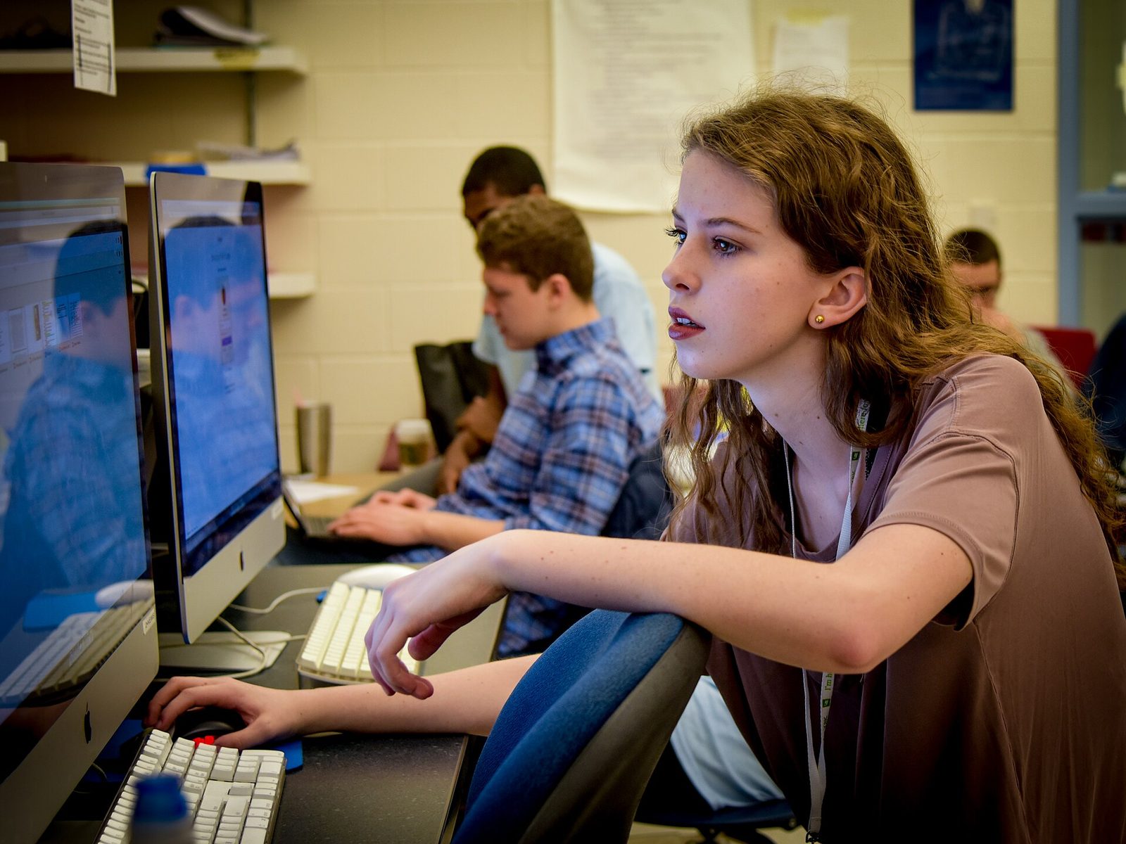 A high school student working at a desktop computer in a classroom lab, with classmates at screens behind her.