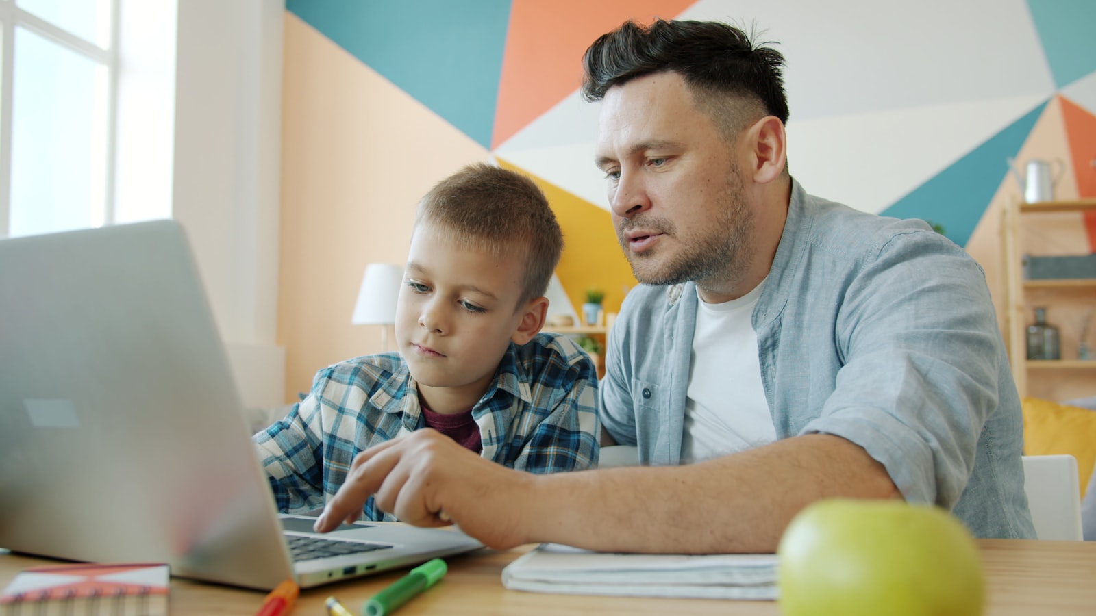 A father sitting beside his son at the kitchen table while the boy works on a laptop and a worksheet.